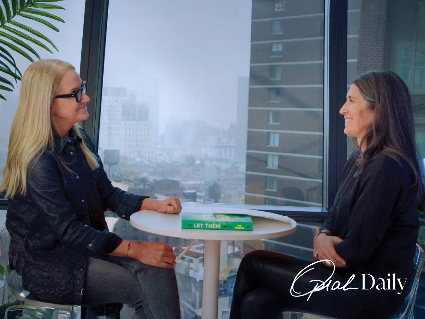 Mel Robbins press image sitting at table with Oprah Daily's editorial director city background